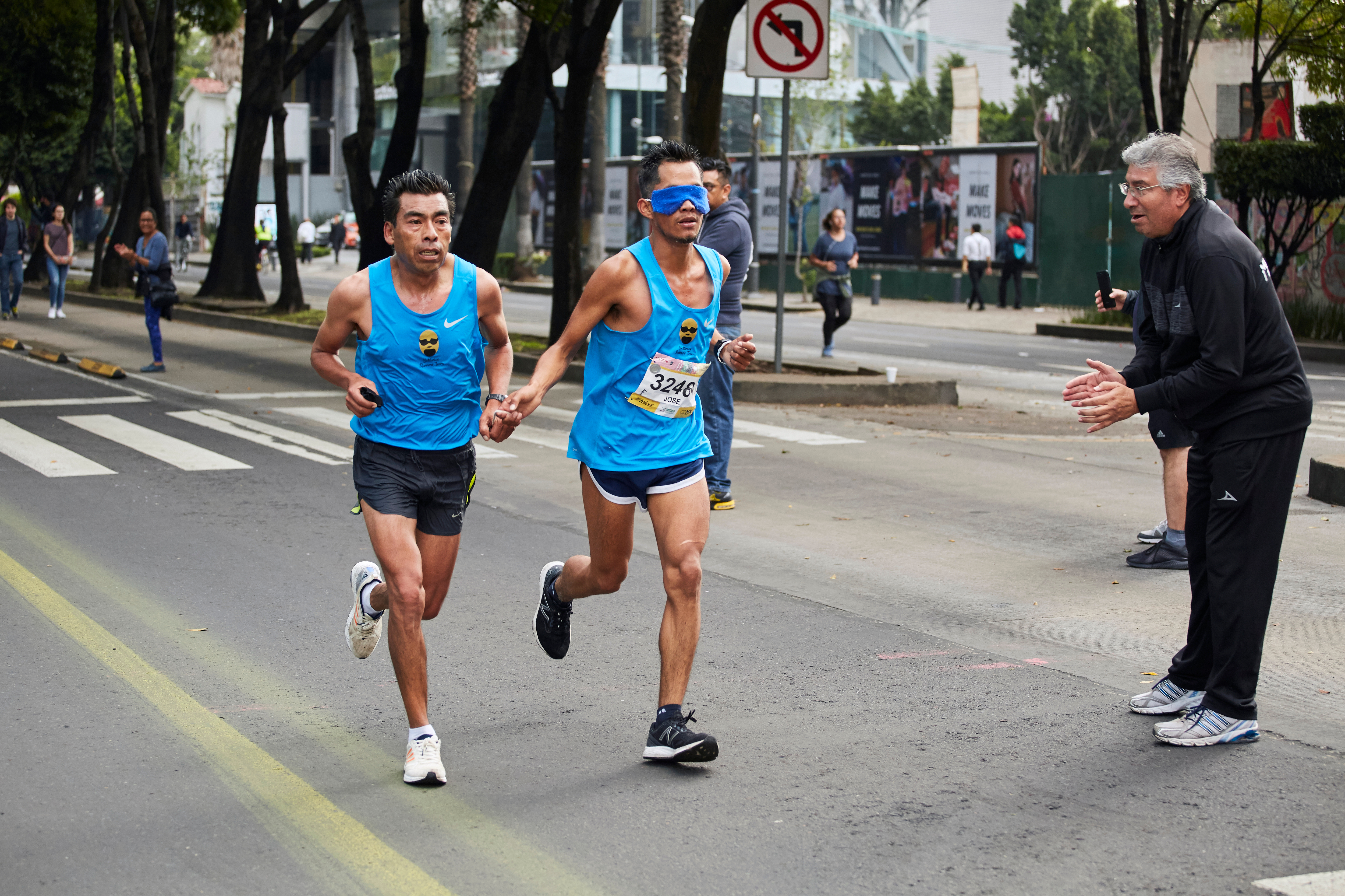 A blindfolded runner holds hands with a sighted guide during a road race, as a coach cheers them on from the sidelines.