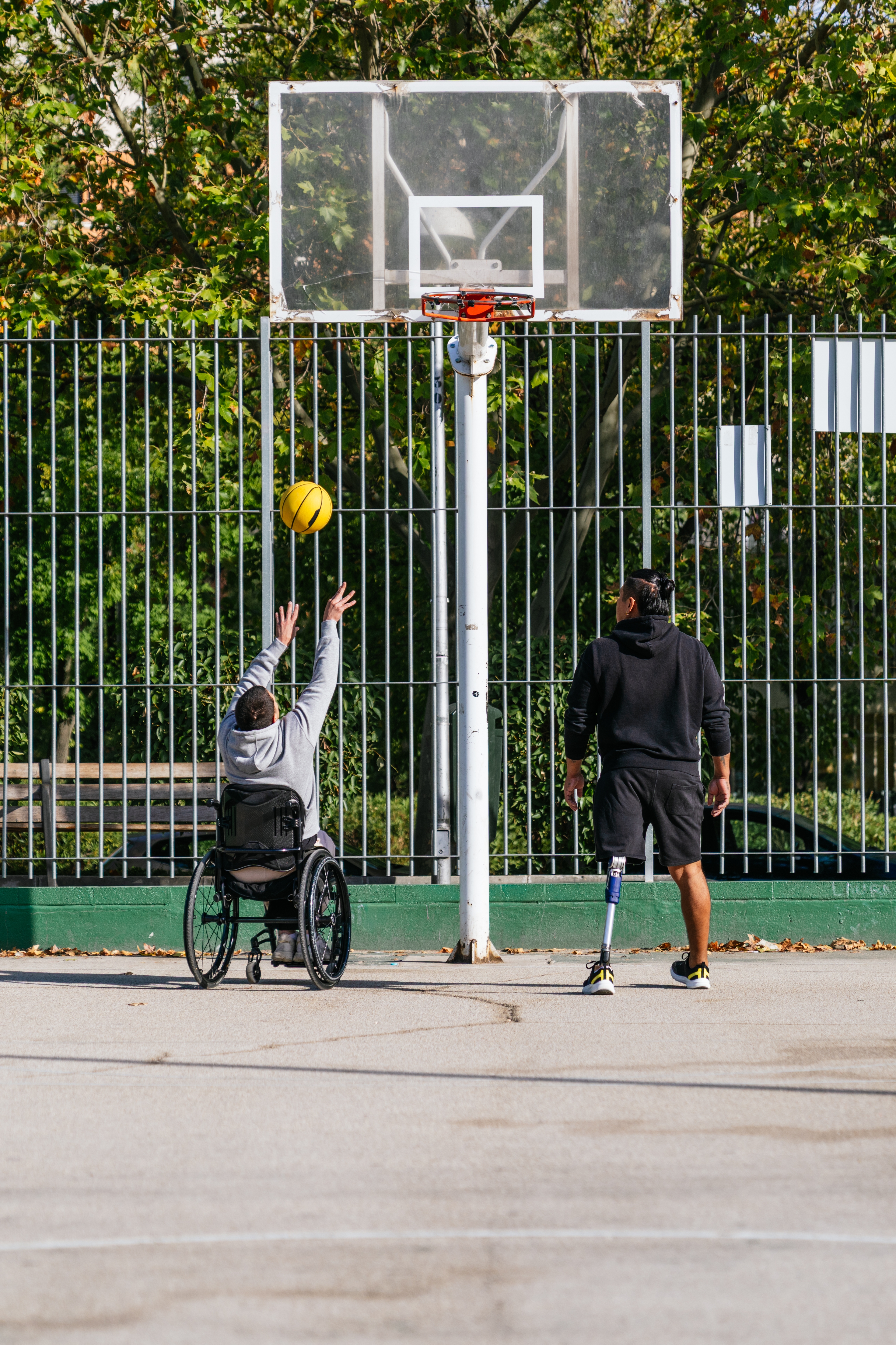 A wheelchair basketball player and a coach with a prosthetic leg practising together on an outdoor court.